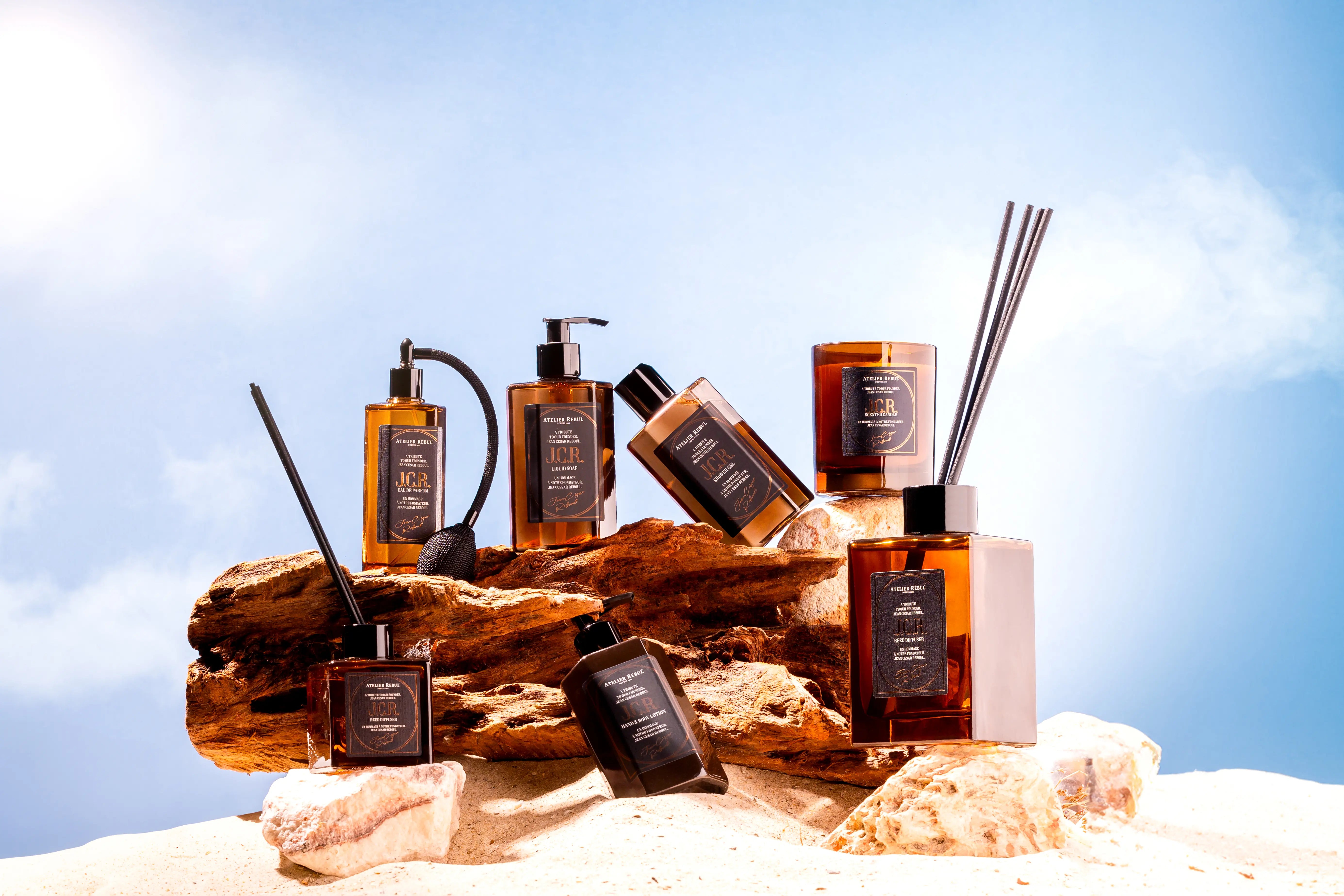 J.C.R. Pflegeserie von Atelier Rebul vor strahlend blauem Himmel – luxuriöse Kollektion mit Seife, Lotion, Parfum, Diffuser und Duftkerze im Bernstein-Design.