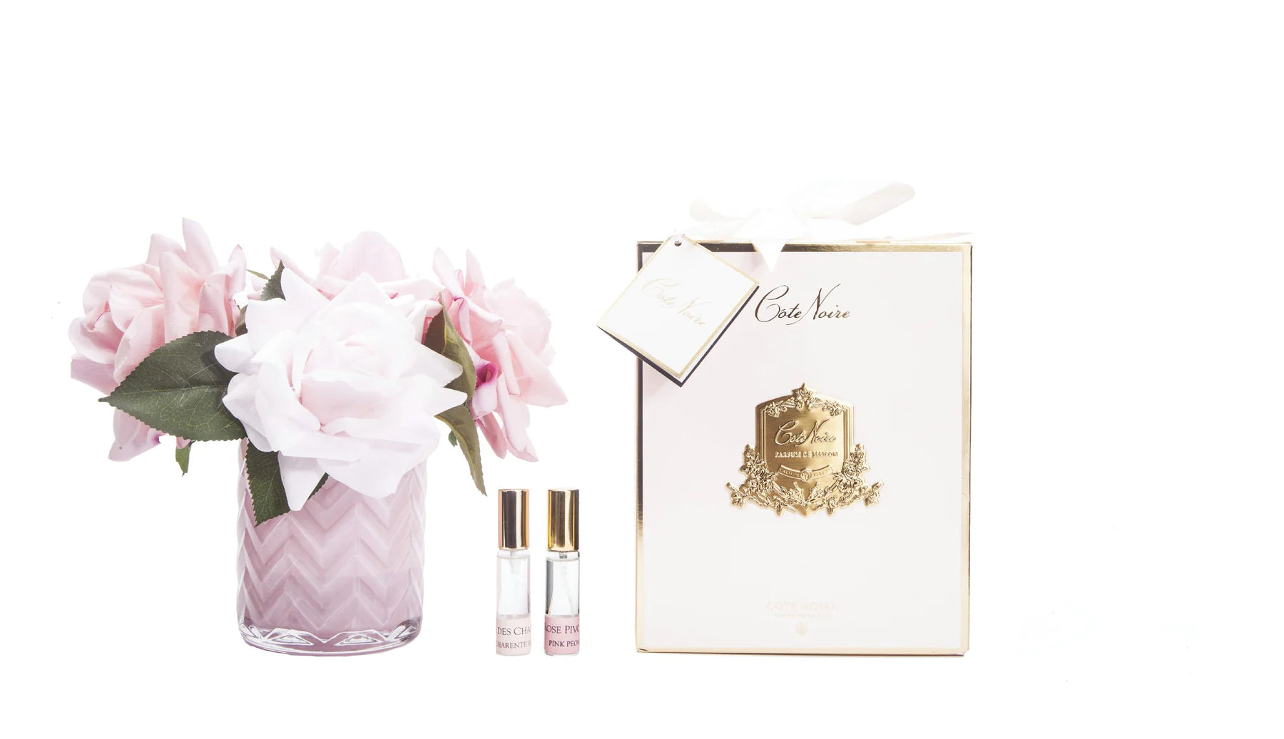 rosa herringbone duftblume in gemustertem glas mit zwei duftsprays und rosa geschenkverpackung vor weissem hintergrund.