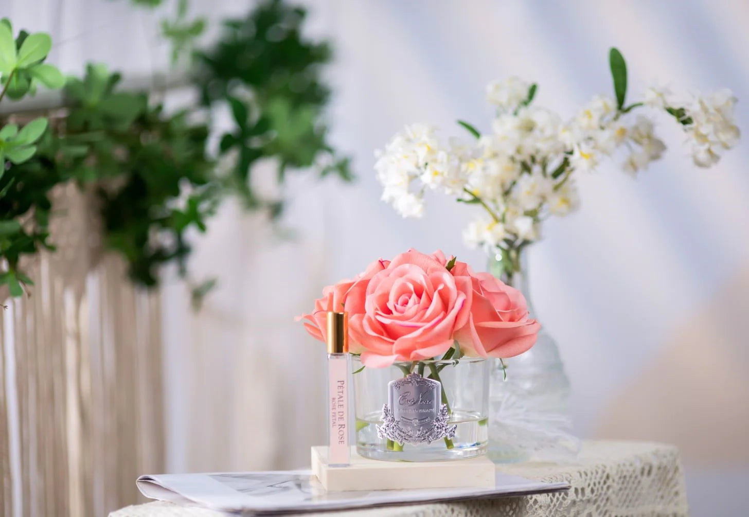 5 rosa duftblumen im glas und duftspray auf weisser unterlage. weitere pflanzen im hintergrund.
