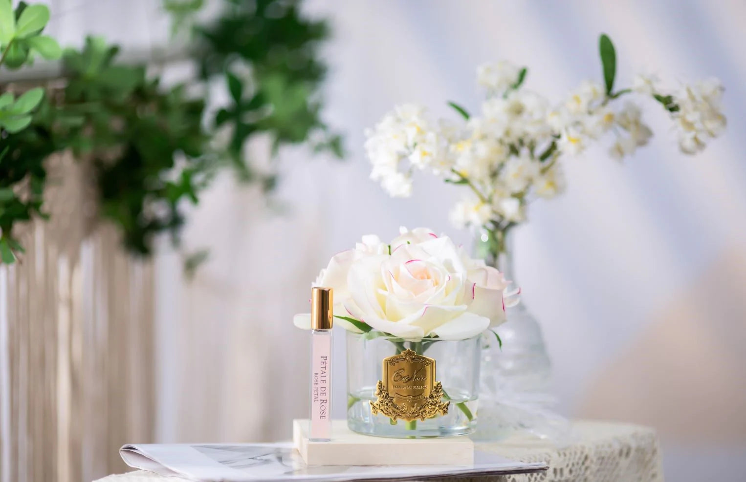 5 rosa duftblumen mit pinkem rand in glas und duftspray auf weißer unterlage. weitere pflanzen im hintergrund.