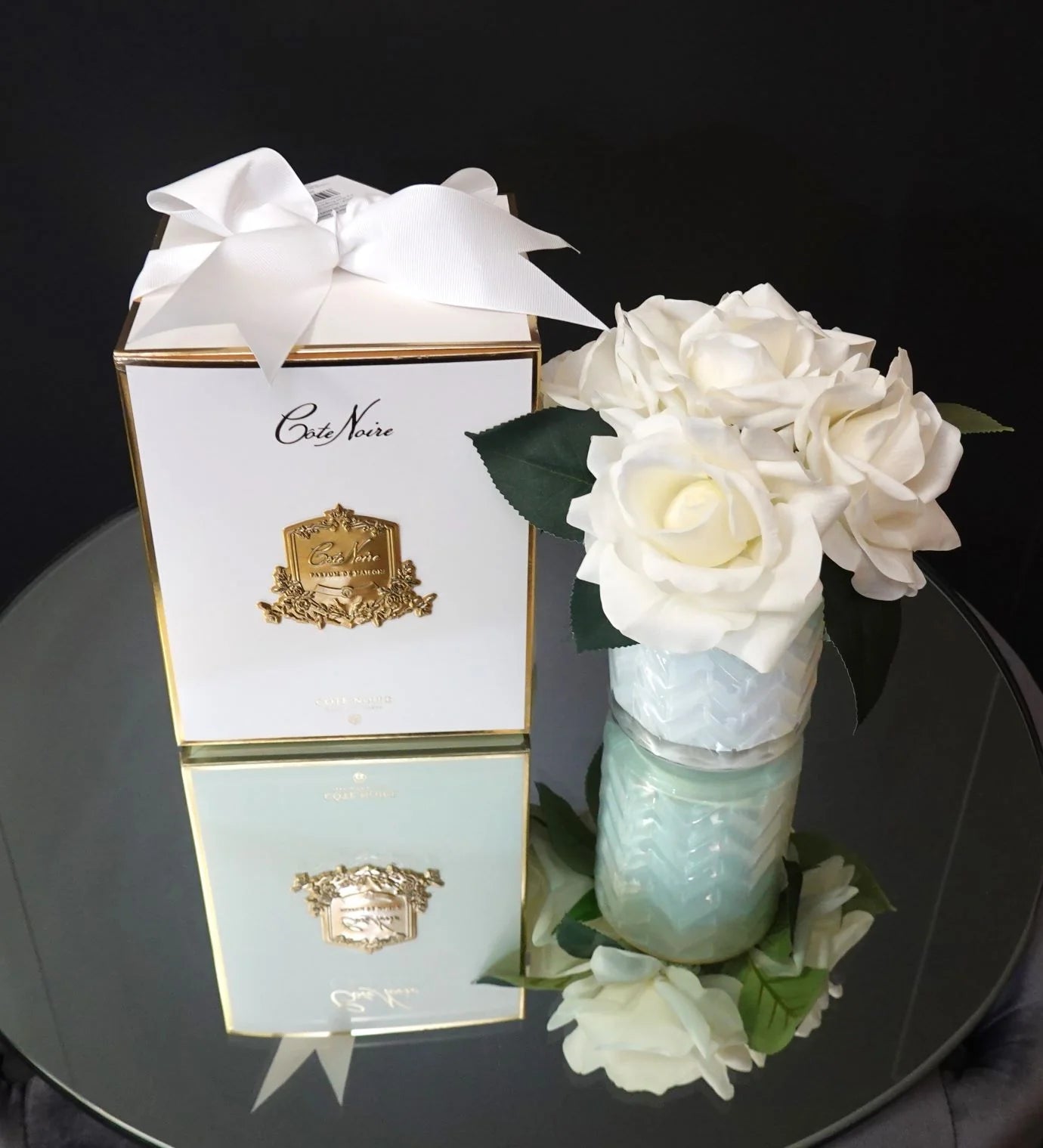 weisse herringbone duftblume neben geschenkbox auf rundem, spiegelndem tisch. obersicht.