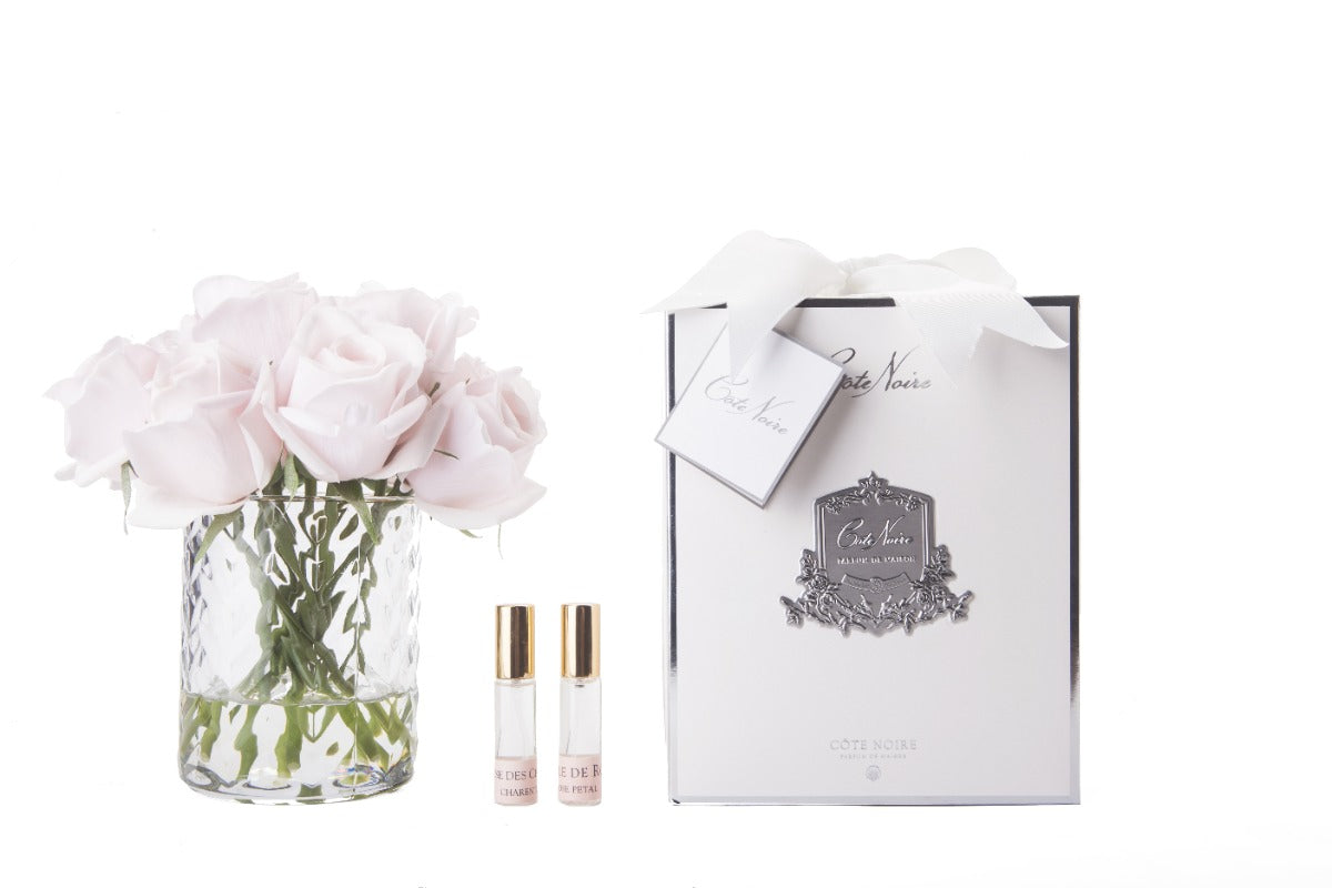 rosa rosenknospen in durchsichtigem glas mit muster. daneben zwei parfumsprays und weisse geschenkbox mit schleife.