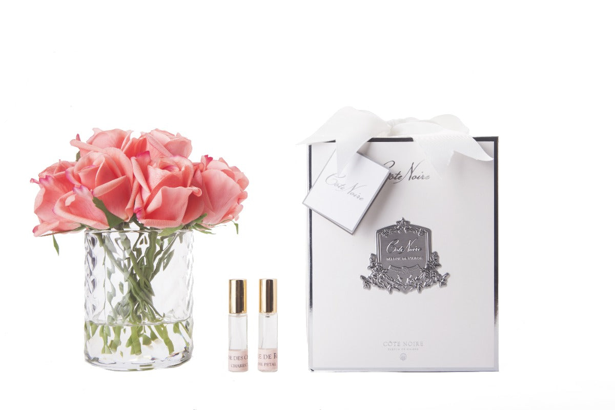 peach rosenknospen in durchsichtigem glas mit muster. daneben zwei parfumsprays und weisse geschenkbox mit schleife.