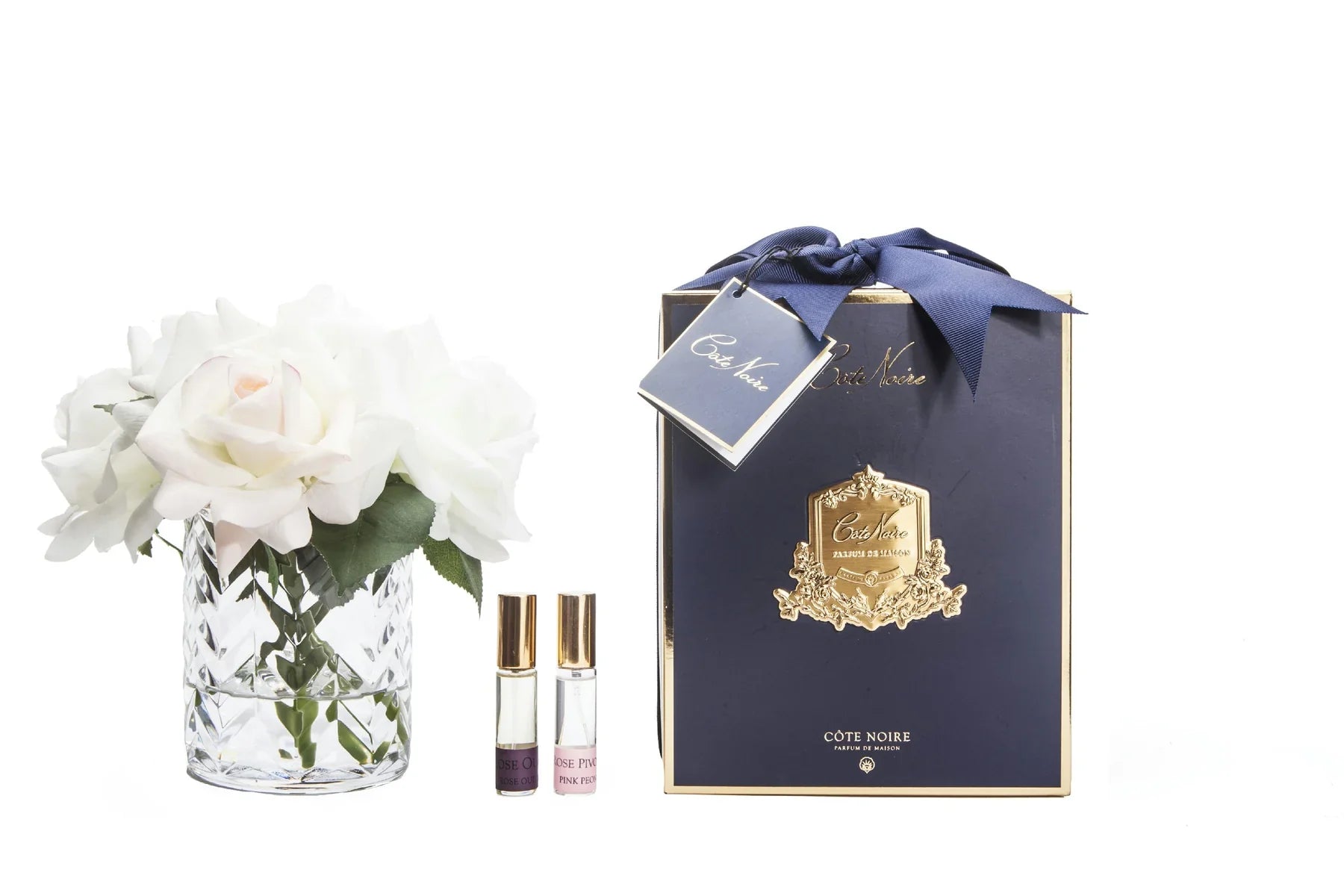 weisse rosen in durchsichtigem glas mit muster. daneben zwei parfumsprays und blaue geschenkbox mit schleife.