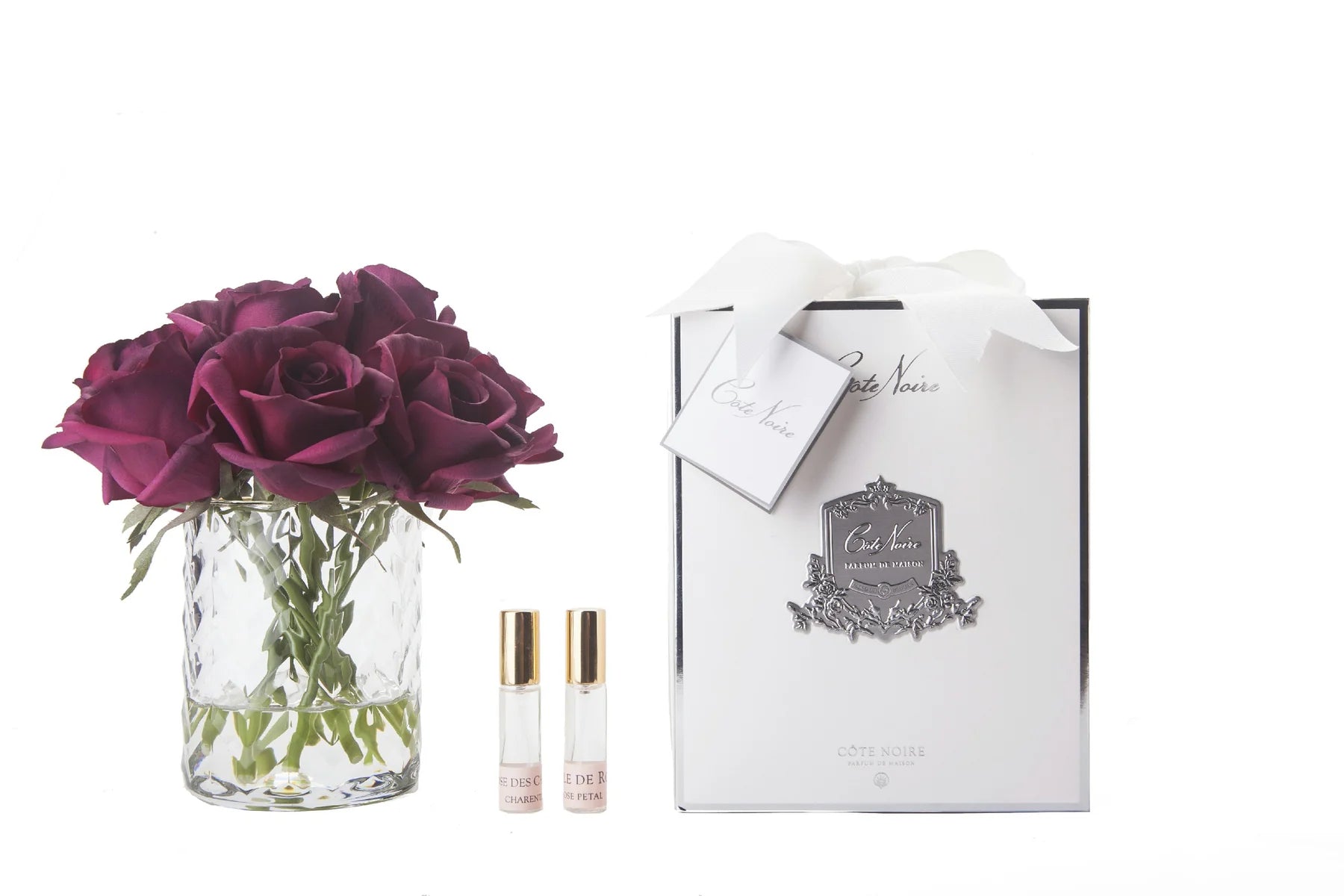 karminrote rosenknospen in durchsichtigem glas mit muster. daneben zwei parfumsprays und weisse geschenkbox mit schleife.