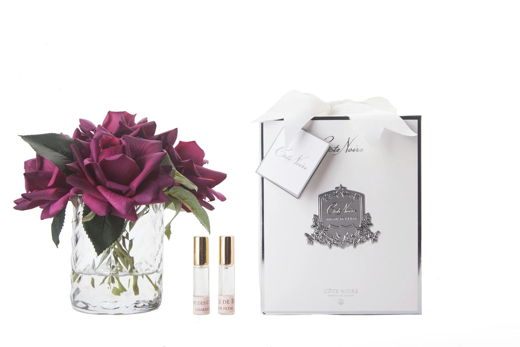 karminrote rosen in durchsichtigem glas mit muster. daneben zwei parfumsprays und weisse geschenkbox mit schleife.