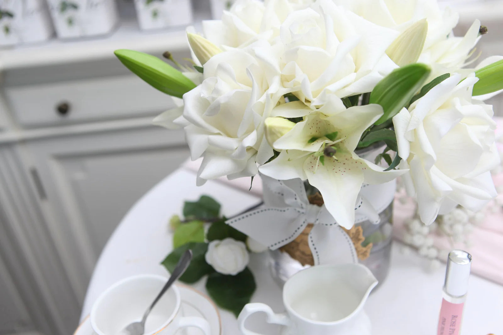 weisse rosen und lilien in hellem glas mit goldenem emblem. weisse schleife um glas gewickelt. set auf esstisch.