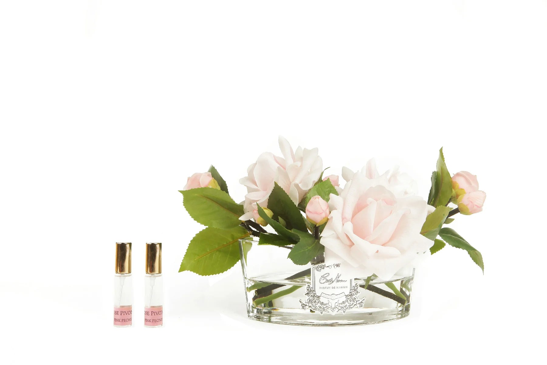 pinke rosen im ovalen glas mit silberner inschrift neben zwei pafrumsprays. weisser hintergrund.