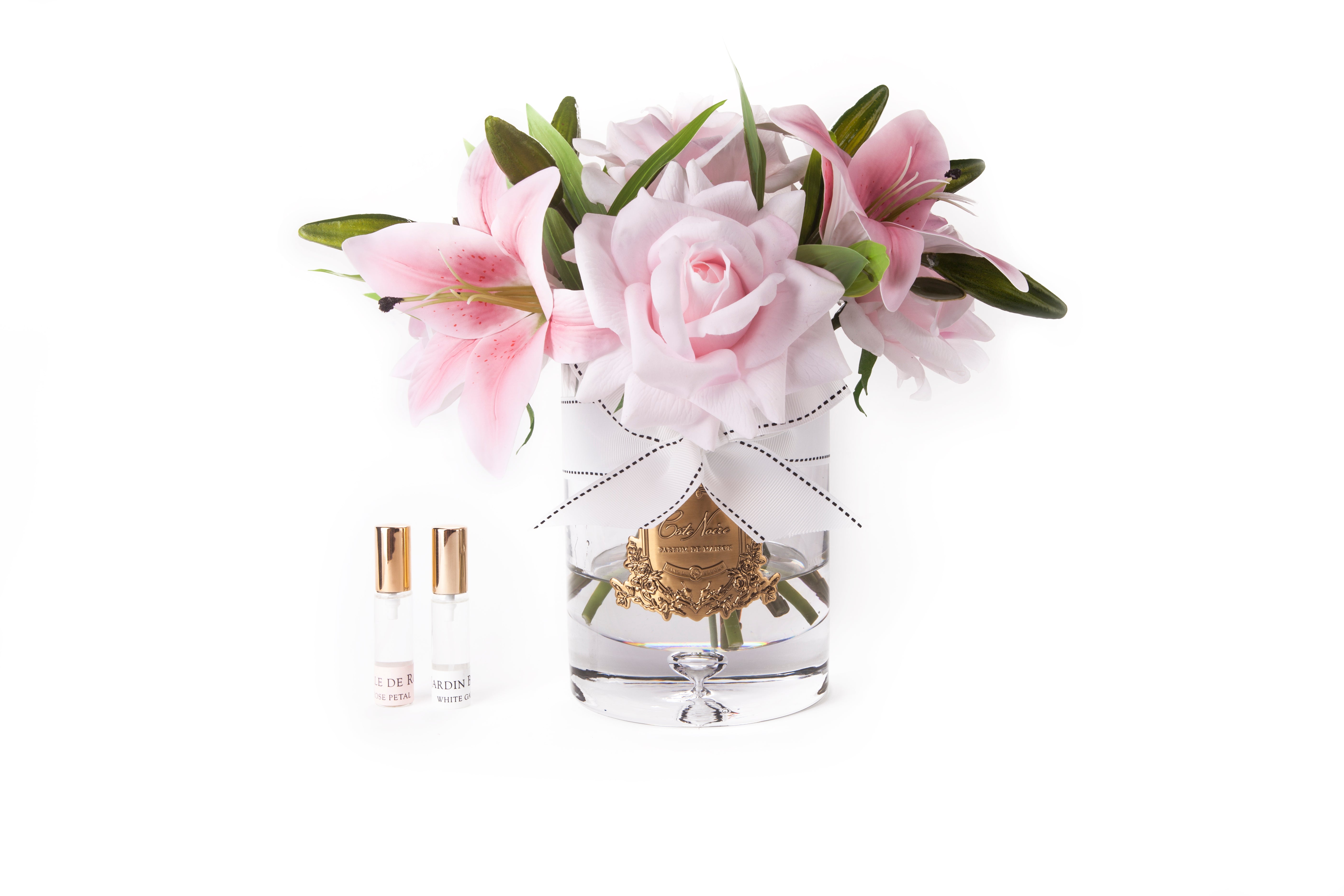 pinke lilien und rosen in hellem glas mit goldener inschrift und weisser schleife. daneben zwei parfumsprays.