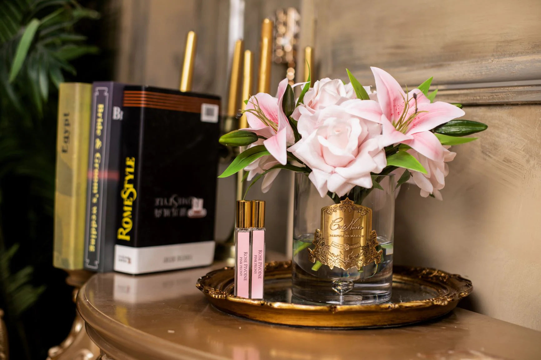 pinke rosen und lilien in hellem glas mit goldenem emblem. daneben zwei duftsprays auf edler unterlage. buecher im hintergrund.