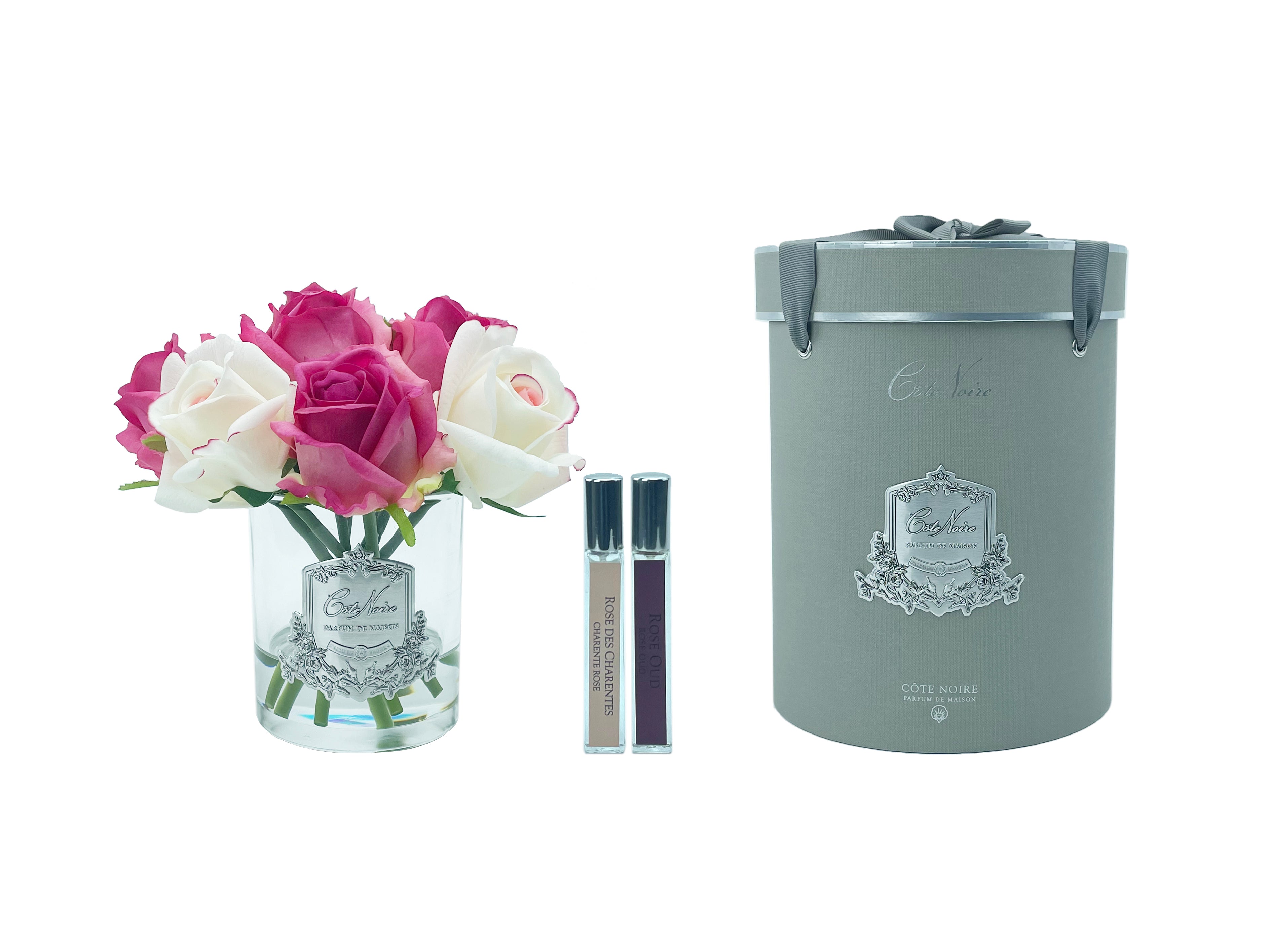 lila und weisse rosen in durchsichtigem glas mit silberner aufschrift, daneben zwei parfumsprays und runde geschenkbox.
