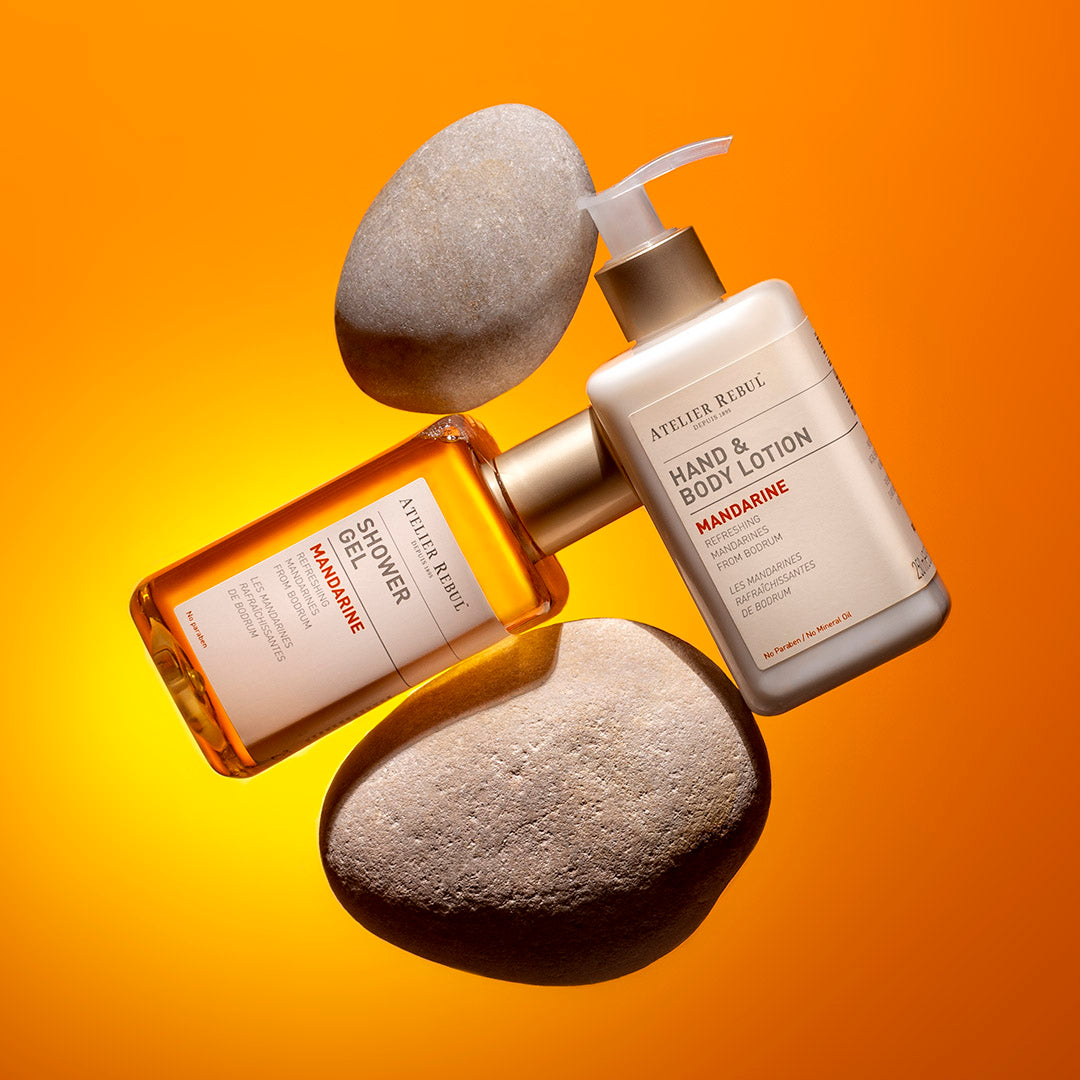 Atelier Rebul Mandarine Shower Gel und Hand & Body Lotion, stilvoll inszeniert mit Steinen vor leuchtend orangefarbenem Hintergrund.