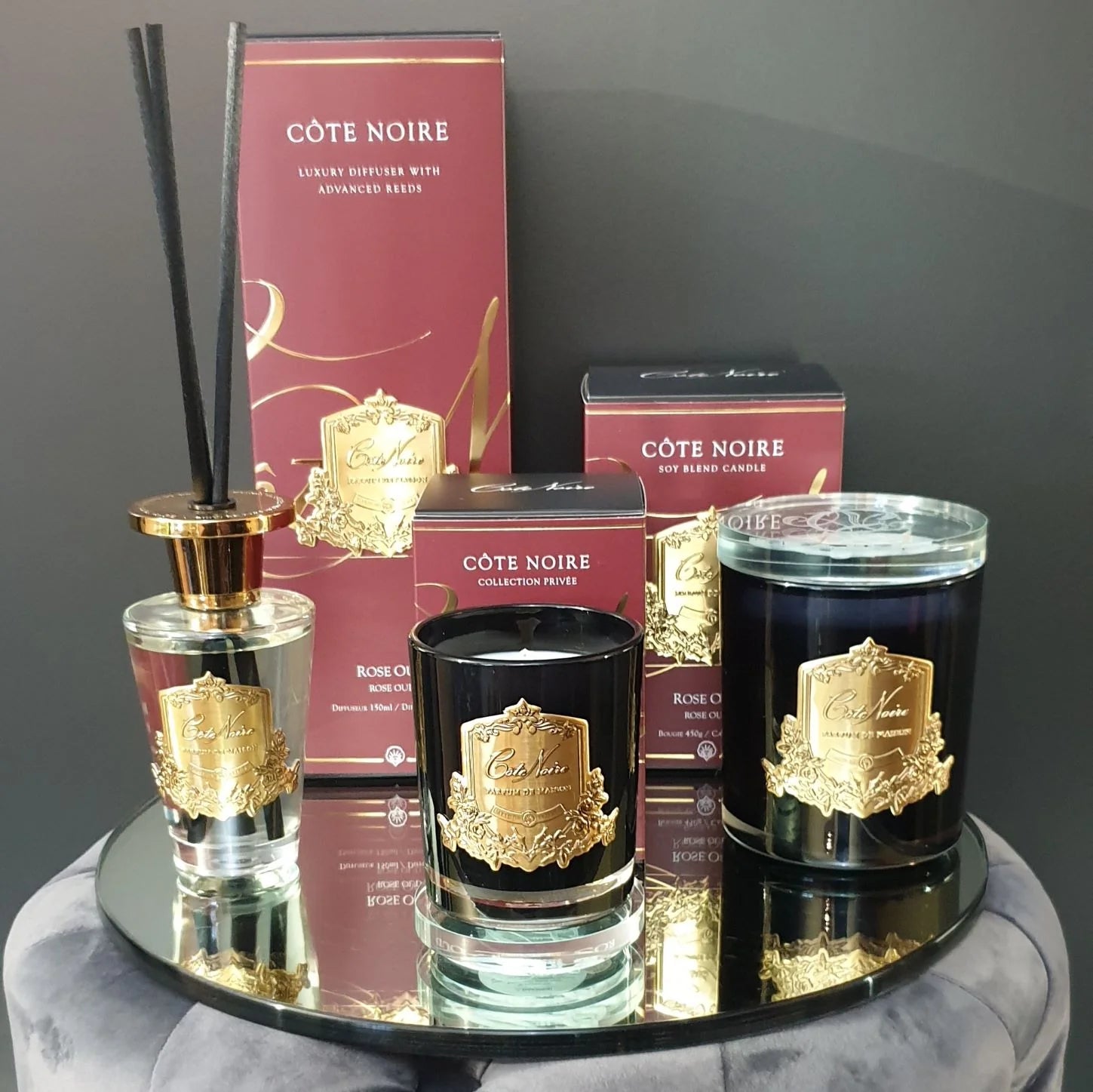 set aus rose oud duftkerzen und raumduft mit dunkelroten verpackungen dahinter auf rundem spiegeltisch.