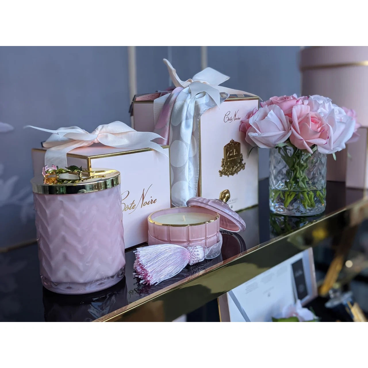 Côte Noire Duftkerze Pink Champagne mit dekorativer Quaste, Geschenkbox und frischen Rosen auf einem Goldregal