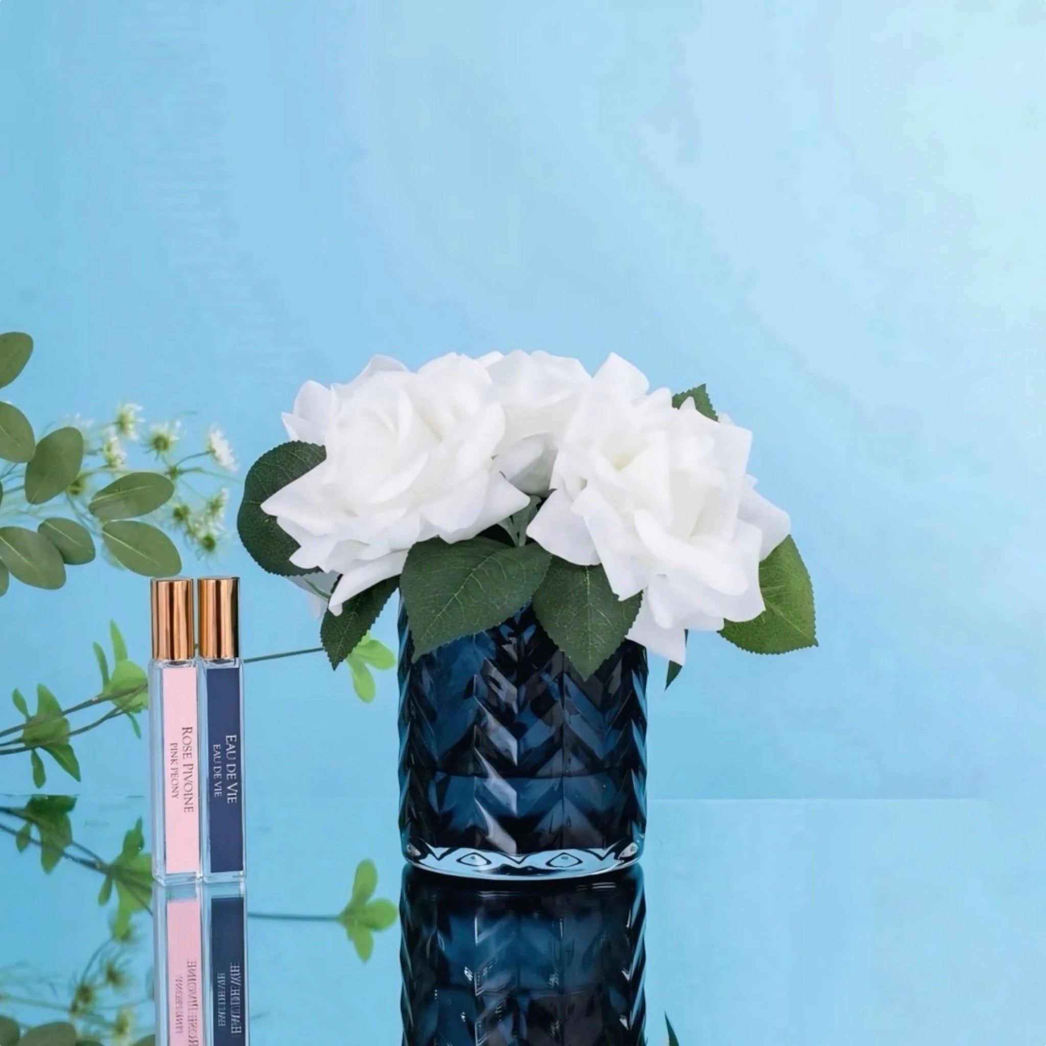 Kuenstliche weiße Rosen in einer blauen Vase neben zwei Parfuemflaschen vor einem hellblauen Hintergrund.