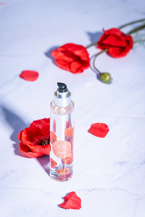runde hohe glasspruehflasche schoene mohnblume steht auf weissem tisch daneben verstreut mohnblumen,