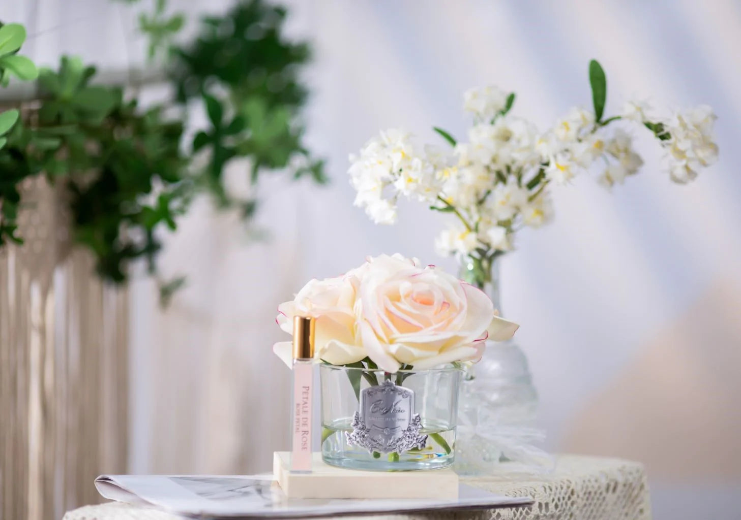 5 weisse duftblumen mit pinkem rand in glas und duftspray auf weisser unterlage. weitere pflanzen im hintergrund.