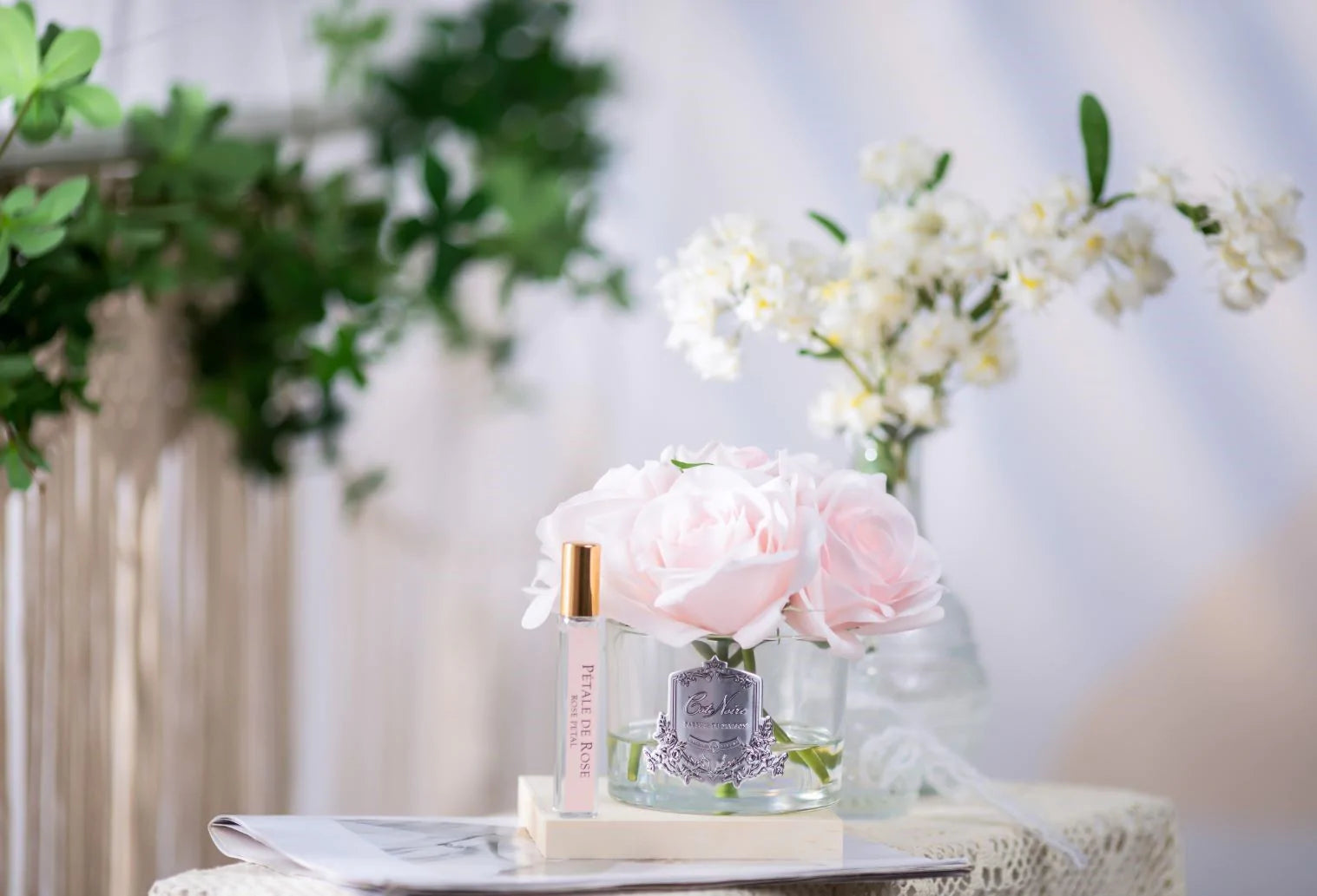 5 pinke duftblumen im glas und duftspray auf weißer unterlage. weitere pflanzen im hintergrund.