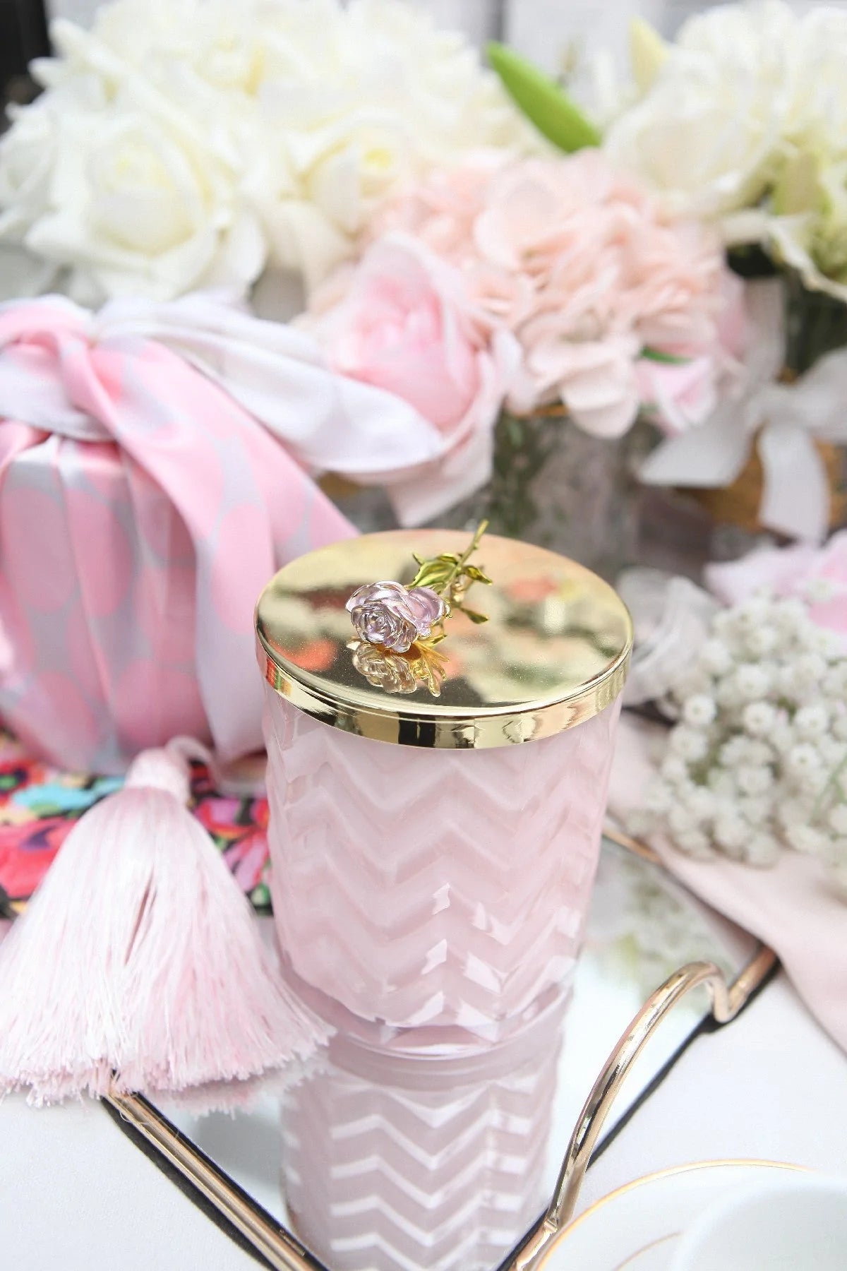 rosa herringbone duftkerze auf einem spiegeltablett, dahinter rosa und weiße duftblumen.