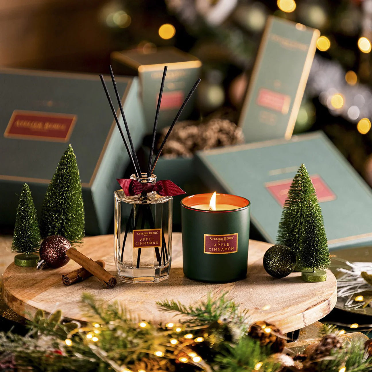 dekor-tannen, sowie frische tannenzweige und gruene geschenkschachteln umgeben die apple cinnamon duftflasche und -kerze.