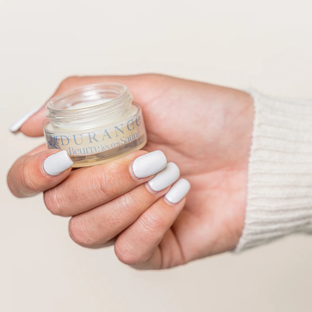 geoeffnete durance lippenbuttertigel liegt in einer frauenhand mit lackierten naegeln hintergrund beige