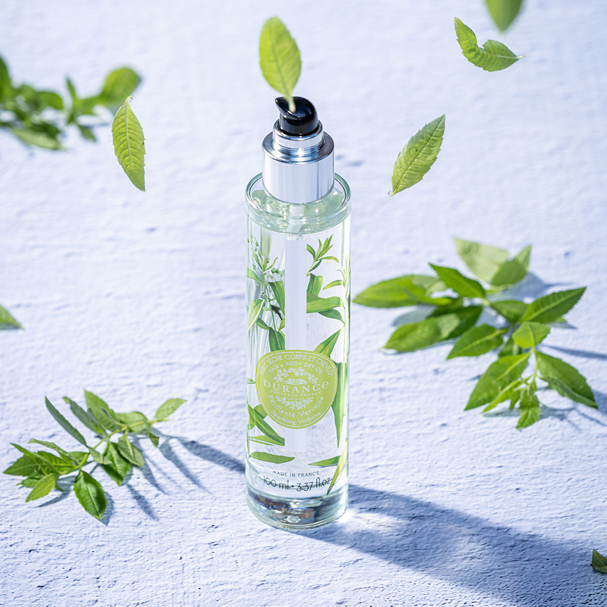runde hohe glasspruehflasche prickelnde verbene steht auf weissem tisch daneben blaetter