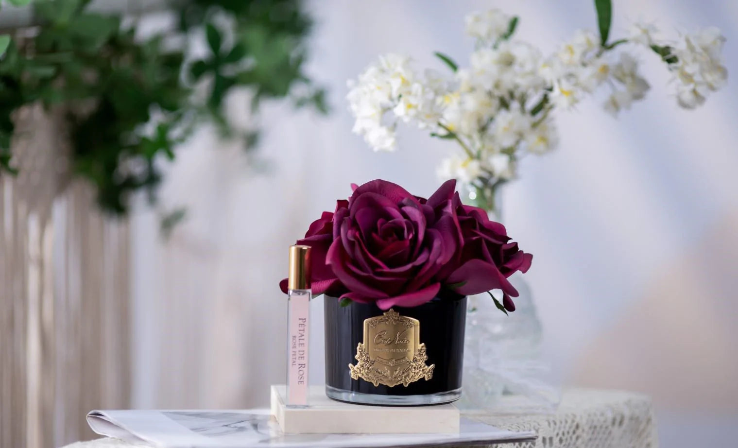 karminrote duftblume in schwarzer vase mit goldenem emblem zusammen mit rose petale duftspray auf einem buch.
