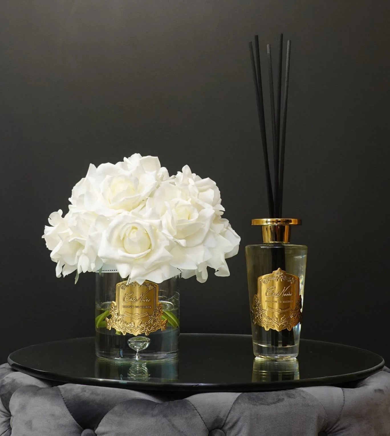 blonde vanilla set aus grand dufststaebchen und duftblume mit weissen rosen auf glastisch.