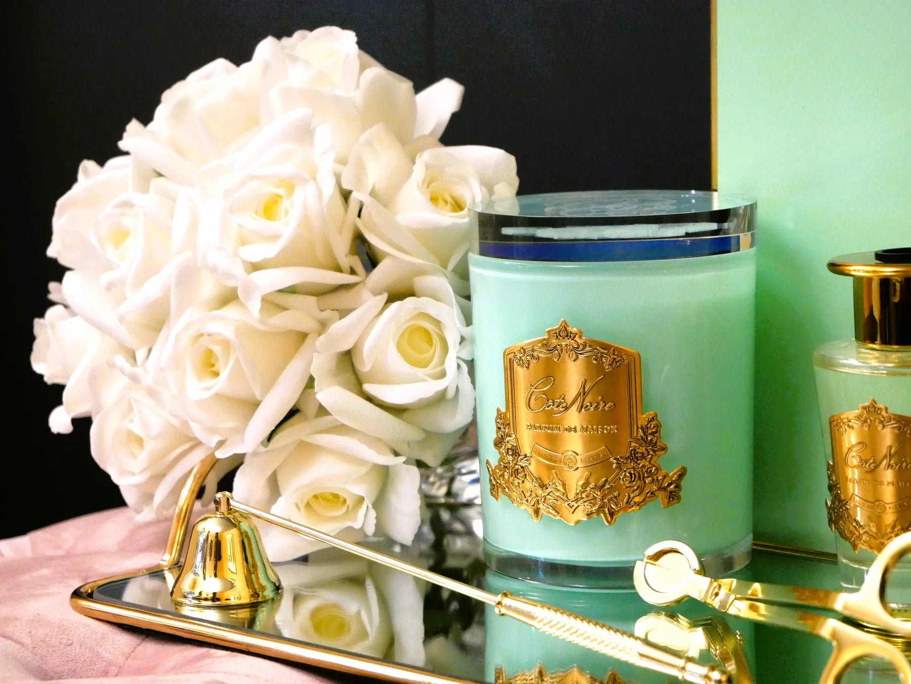duftkerze in jade gruenem glas mit goldener inschrift und glasdeckel neben weissen rosen vor schwarzem hintergrund.