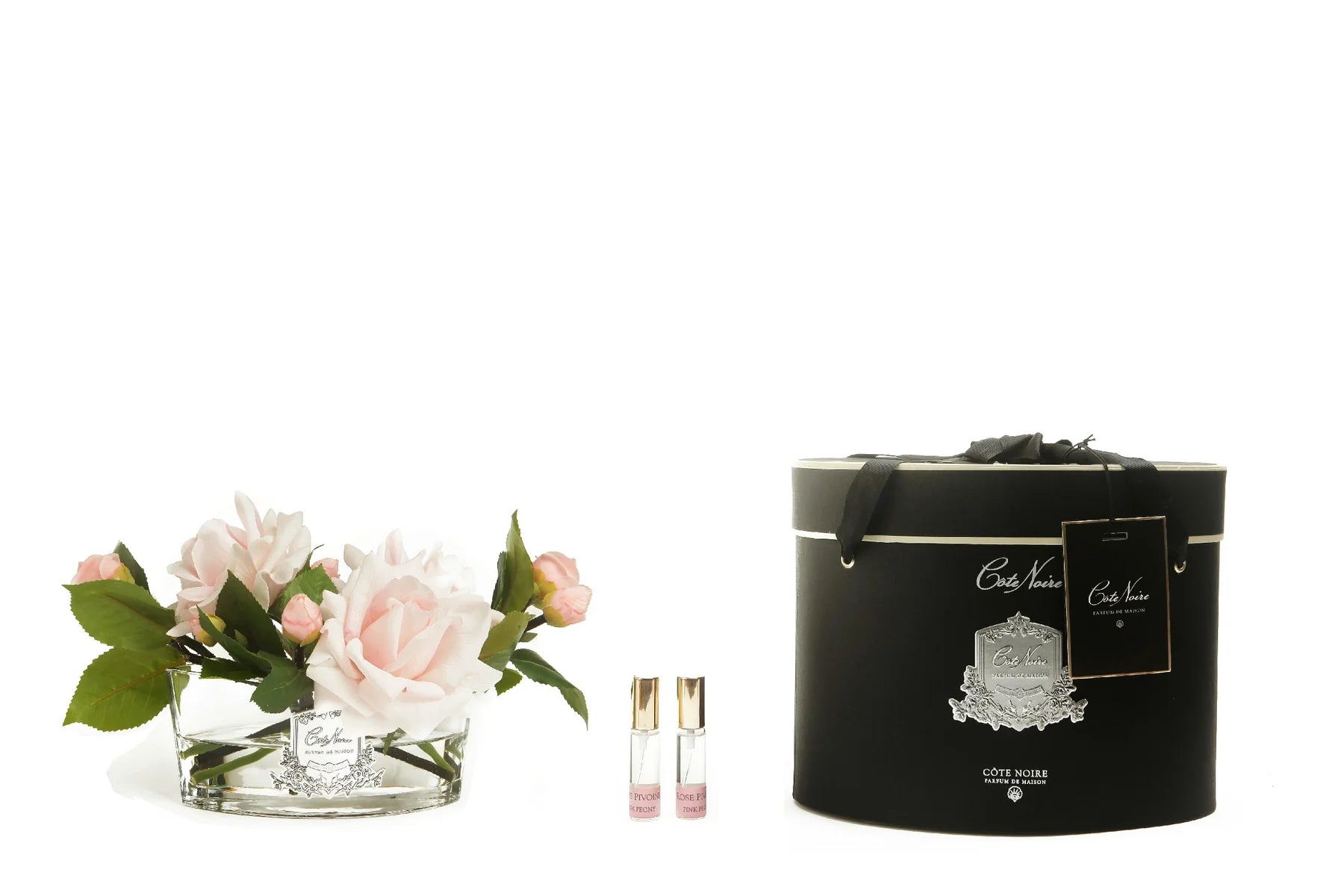 pinke rosen in hellem, ovalem glas mit silberner inschrift. daneben zwei parfumsprays und runde geschenkbox.