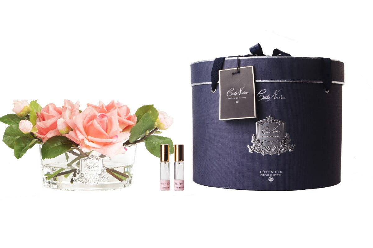 pfirsichfarbene rosen in hellem, ovalem glas mit silberner inschrift. daneben zwei parfumsprays und runde geschenkbox.