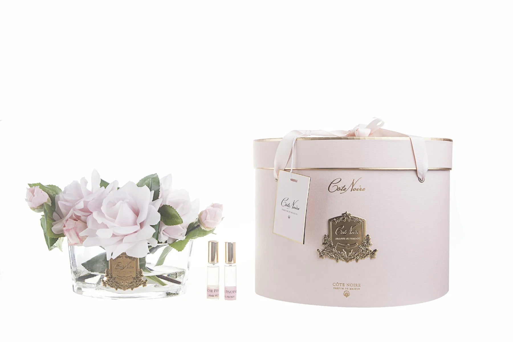 pinke rosen in hellem, ovalem glas mit goldener inschrift. daneben zwei parfumsprays und runde geschenkbox.