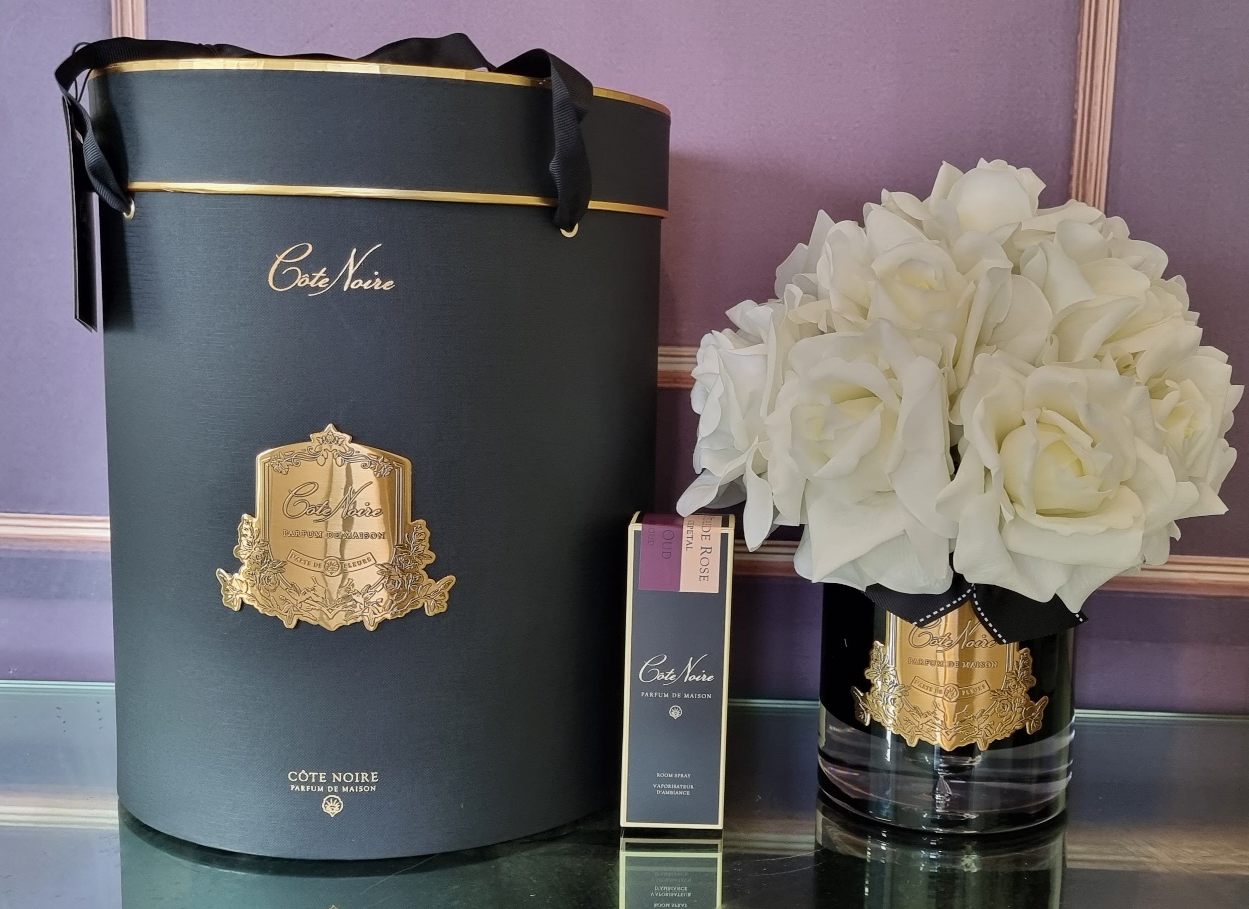 weisse duftblumen in schwarzem glas mit goldener inschrift neben schwarzer geschenkbox.