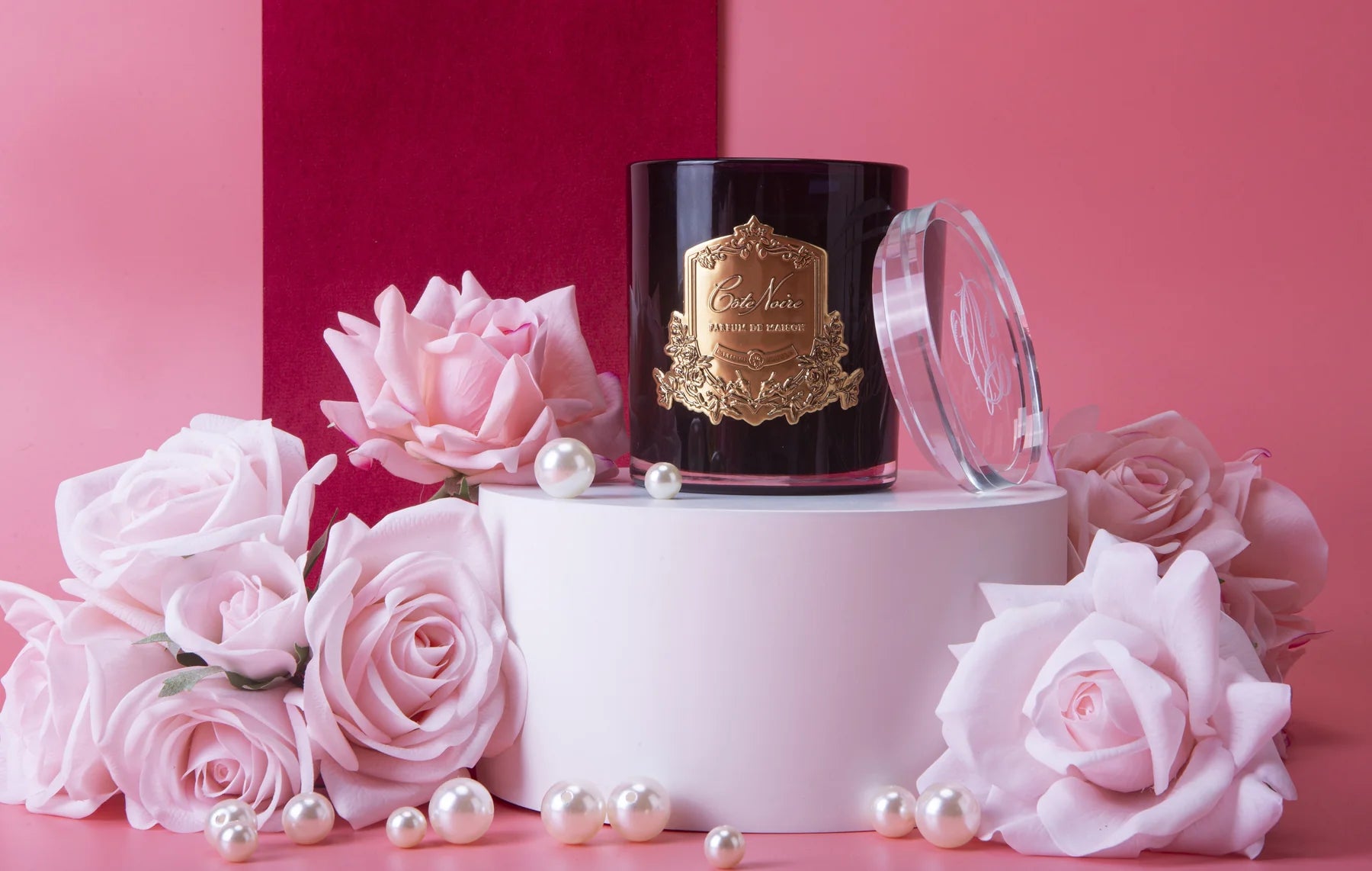 rose petal duftkerze in dunklem glas mit goldener inschrift auf pinkem podest, umgeben von pinken rosen. pinker hintergrund