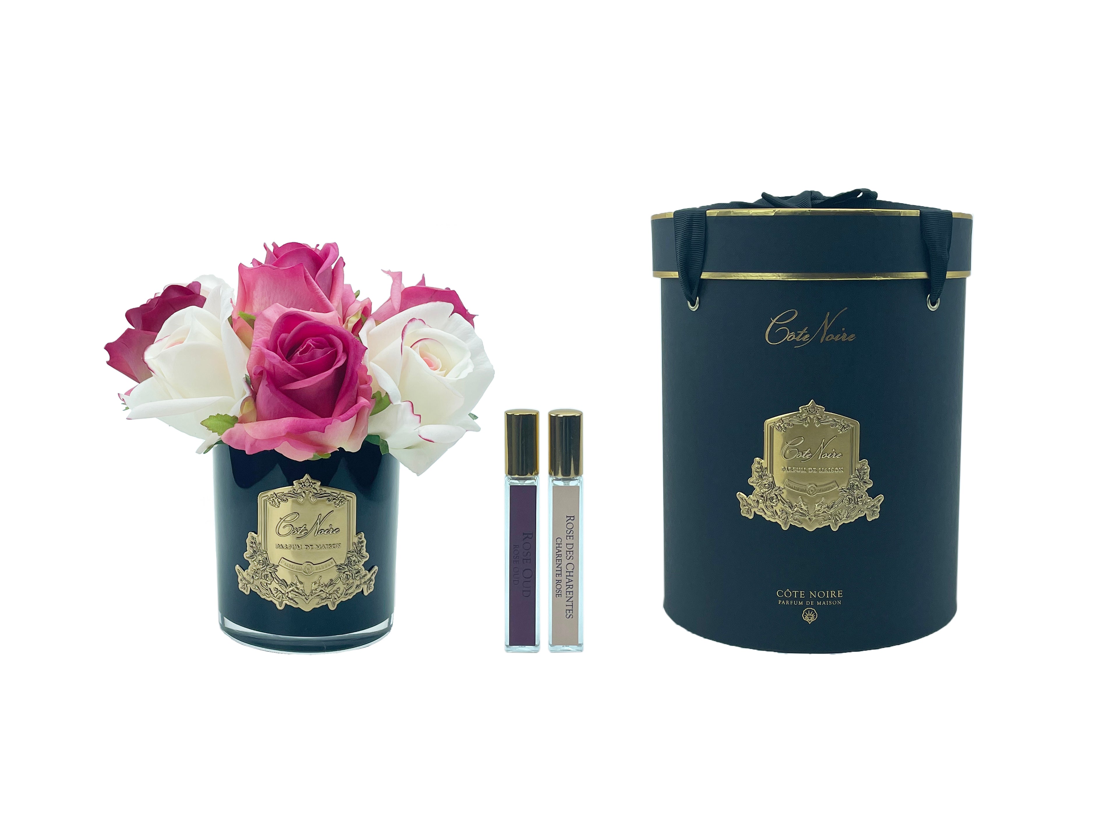 lila und weisse rosen in schwarzem glas mit goldener aufschrift, daneben zwei parfumsprays und runde geschenkbox.