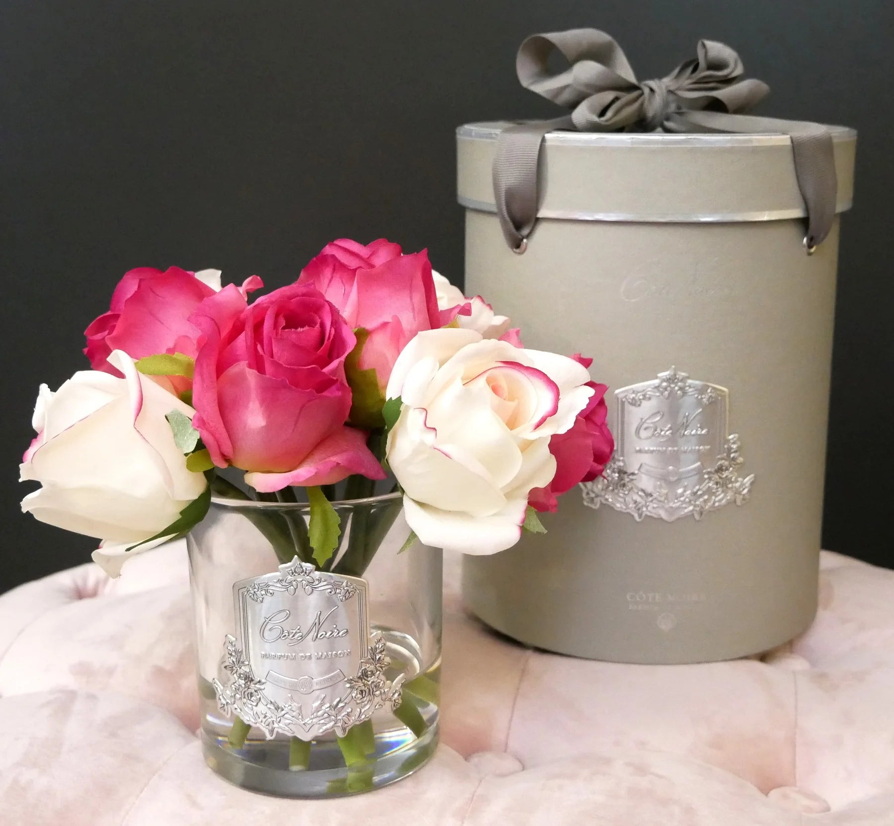 pink und weiss gemischter rosenstrauss in hellem glas vor zylinderfoermiger, silberner geschenkbox