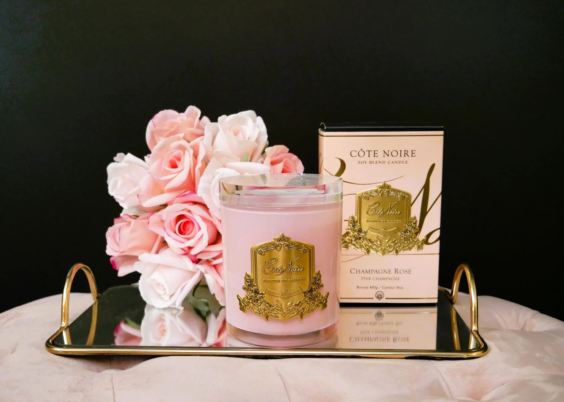pinke duftkerze mit goldener inschrift, dahinter verpackung und duftblumen in pink auf goldenem serviertablett. frontansicht