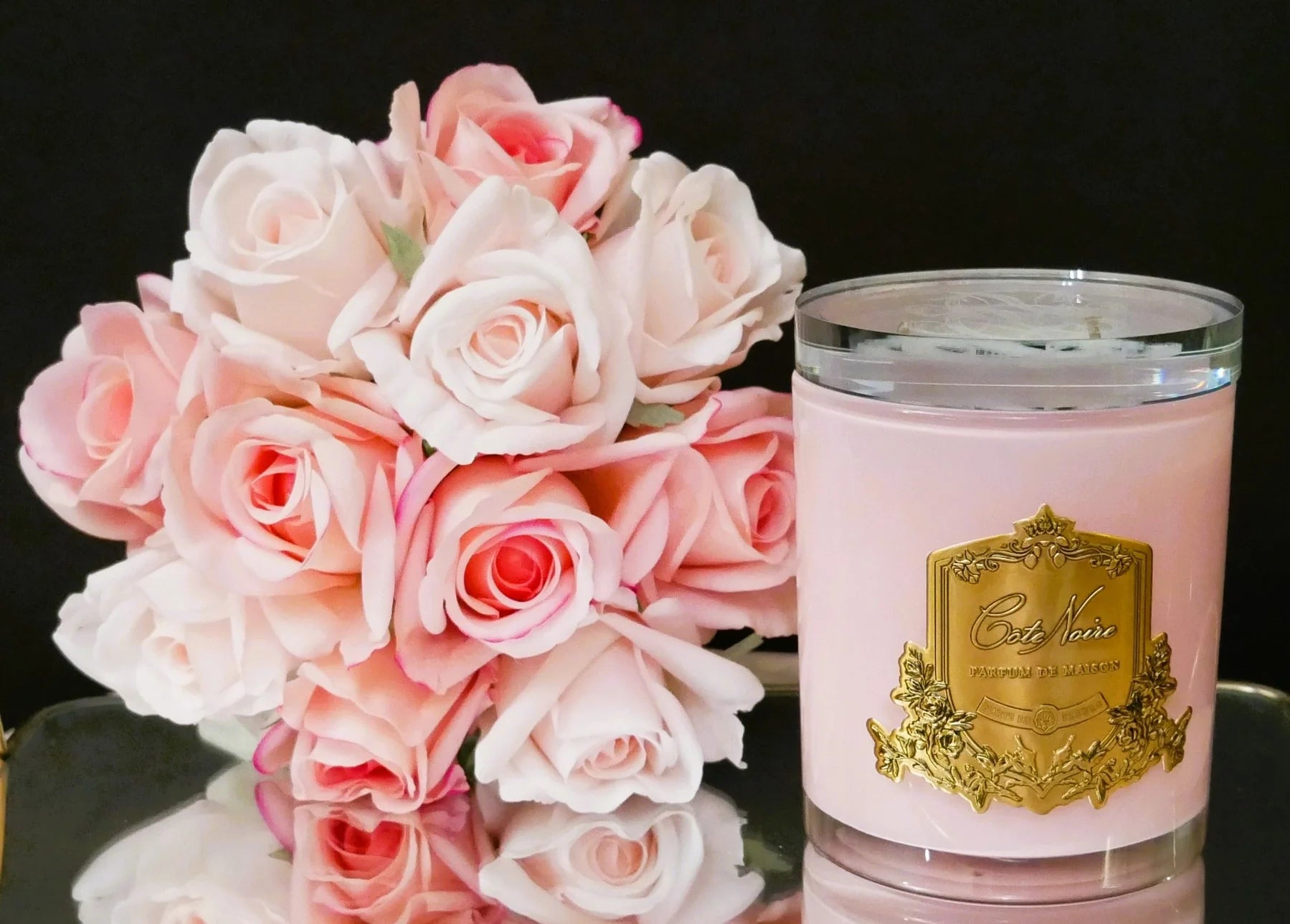 duftkerze in pinkem glas mit goldener inschrift und glasdeckel neben pinken und weissen rosen vor schwarzem hintergrund.