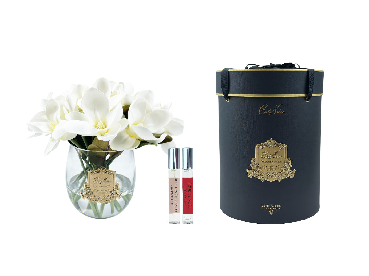 weisse magnoliasblueten in gerundeter glasvase neben duftsprays und dunkler runder geschenkbox.