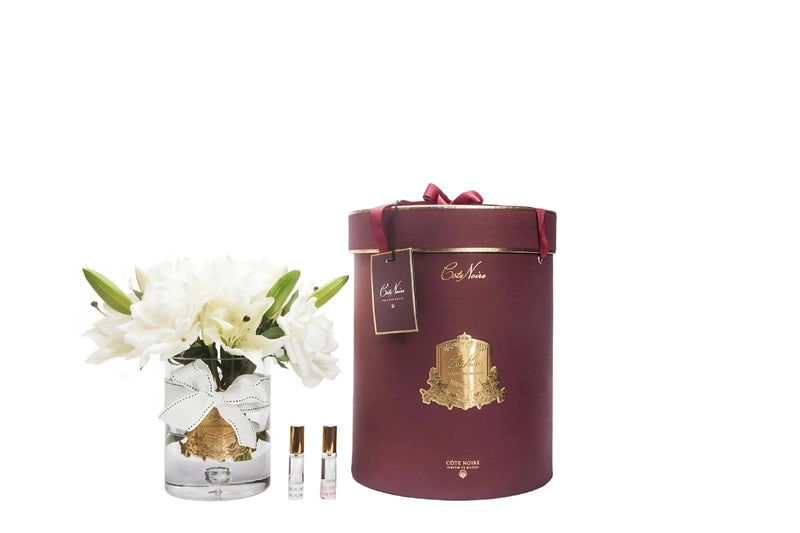 weisse luxury lilies duftrosen im glas neben burgunderfarbener zylenderbox. dazwischen zwei parfumsprays.