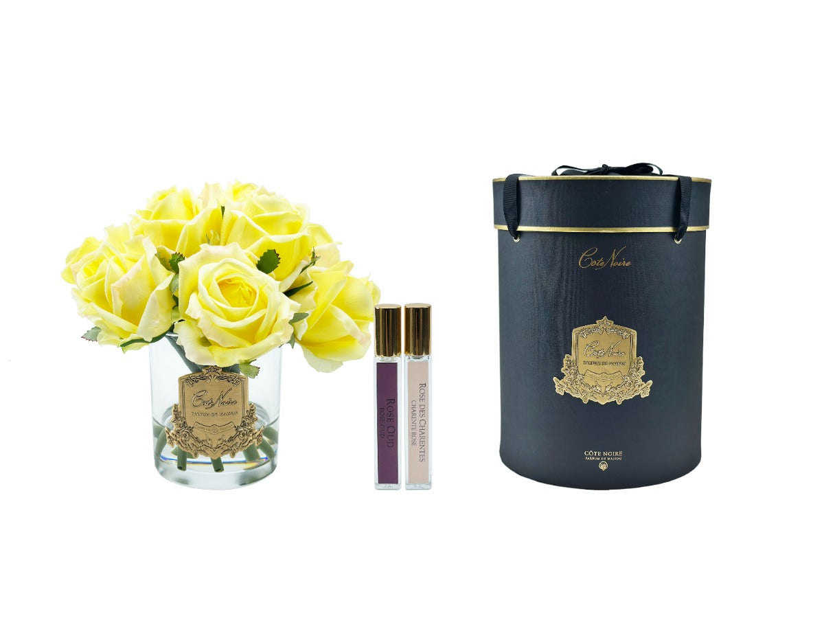 gelbe rosen in durchsichtigem glas mit silberner aufschrift, daneben zwei parfumsprays und runde geschenkbox.