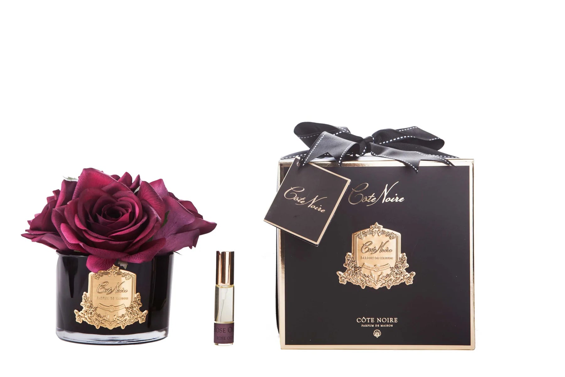karminrote rosen in schwarzem glas mit goldenem emblem, daneben das duftspray und eine geschenkverpackung mit schleife.