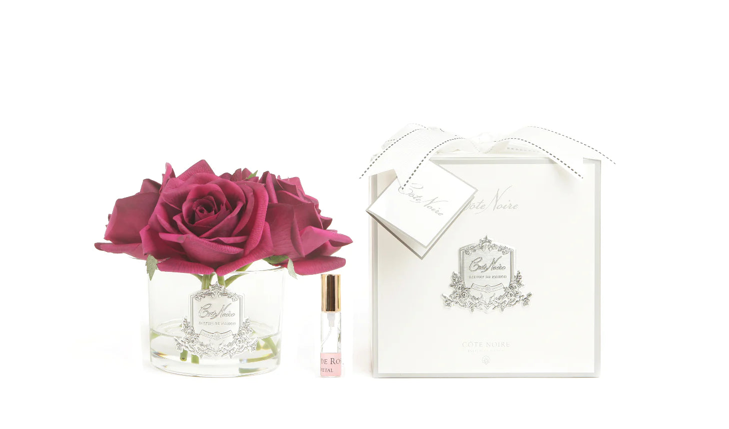 5 karminrote rosen mit in hellem glas mit silbernem emblem. dazu duftspray und weisse verpackung. weisser hintergrund.