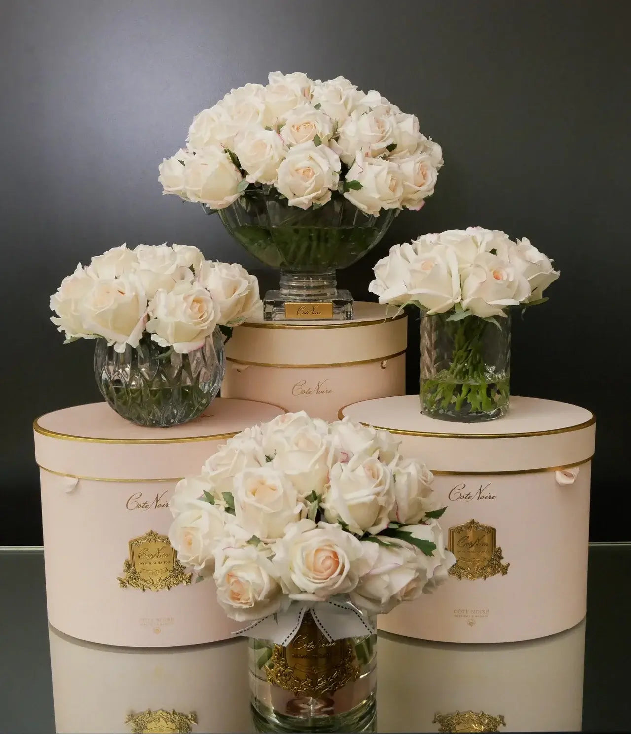 Vertikales Arrangement mit Blush Duftblumen – Kombination aus runder, kristallklarer und goldverzierter Vase auf rosa Boxen
