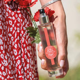 weibliche hand haelt runde glaspruehflasche eau fraiche schoene mohnbluete mit 2 mohnblueten seitlich noch ein stueck vom roten kleid