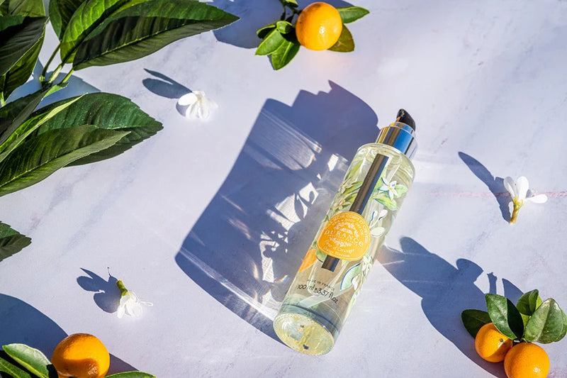 runde hohe glasspruehflasche orangenbluete liegt auf weissem tisch daneben manderinen, blueten und blaetter