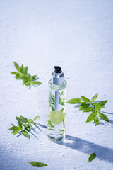 runde hohe glasspruehflasche prickelnde verbene steht auf weissem tisch daneben blaetter