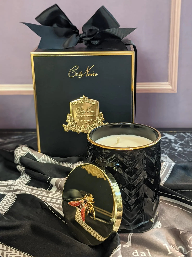 herringbone duftkerze mit angelehntem golddeckel auf schwarzem seidenschal. dahinter die geschenkbox.
