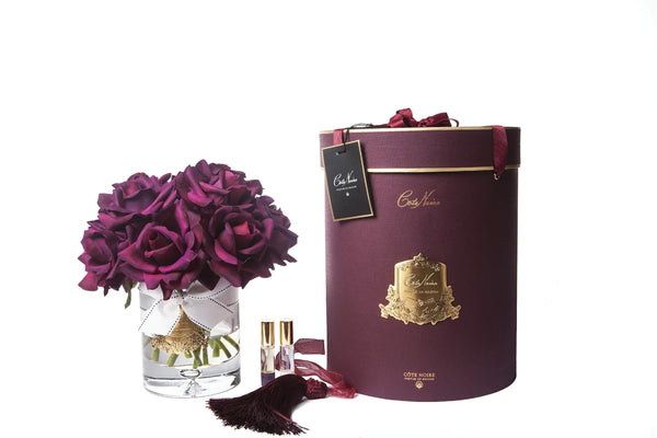 13 karminrote rosen arrangiert in einer edlen glasvase, zwei parfumsprays und eine luxurioese runde geschenkbox in burgunder.