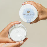 2 frauenhaende halten den durance balsam tigel sowie den verschluss hintergrund ist beige